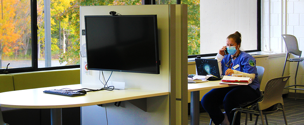 A student sits at a desk using a laptop to study. On the facing side, a widescreen monitor is connected to a keyboard and mouse